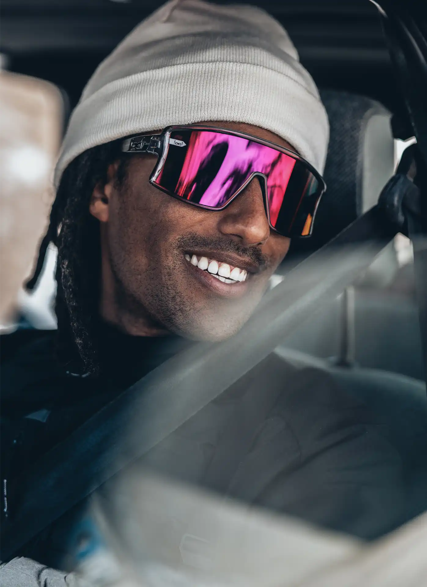 Ein Mann mit weißer Beanie und verspiegelter Sport-Sonnenbrille mit pinken Gläsern sitzt im Auto und lächelt breit. Er trägt einen Sicherheitsgurt, und das Licht spiegelt sich auf der Fensterscheibe. Die Szene wirkt entspannt und urban.